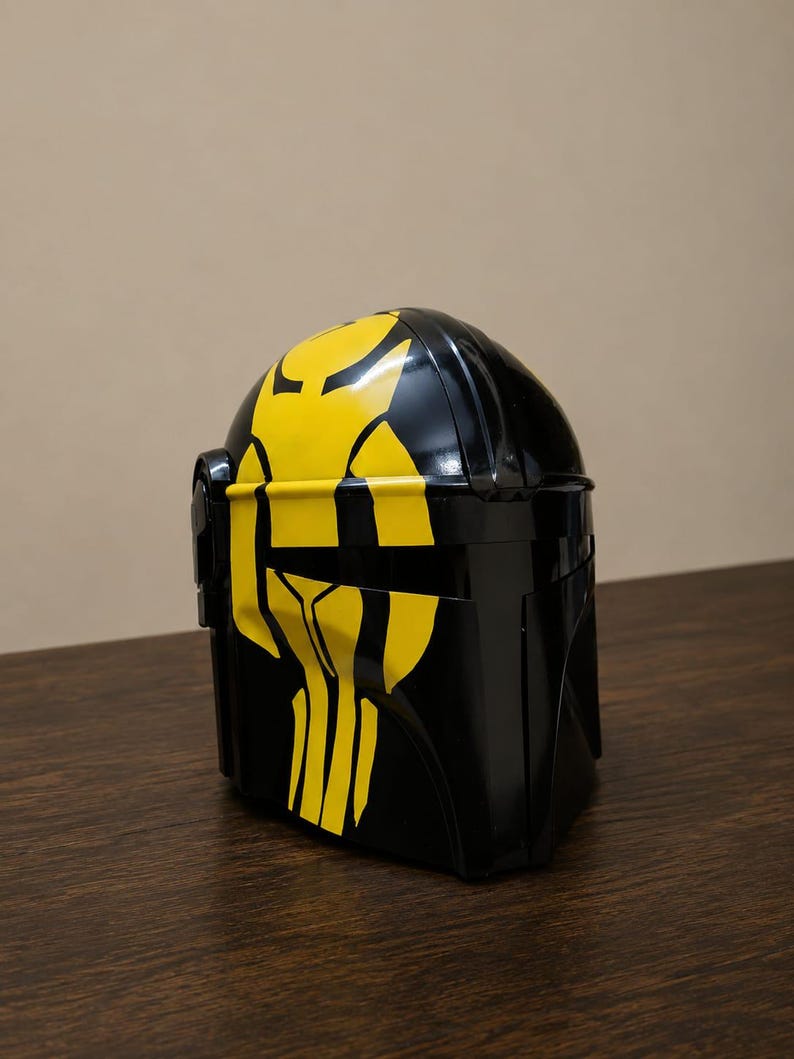 May include: A glossy black helmet featuring a prominent yellow graphic design on the front. The helmet has a smooth, curved surface with a horizontal visor opening and a distinct, angular shape that covers the head and face area completely.