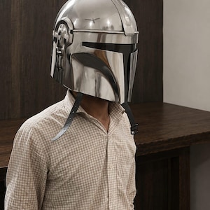 May include: A polished silver-colored metal helmet with a smooth, reflective surface and a T-shaped black visor opening. The helmet features a central ridge, side ear-cap details, and two thin black leather chin straps hanging down from the sides.