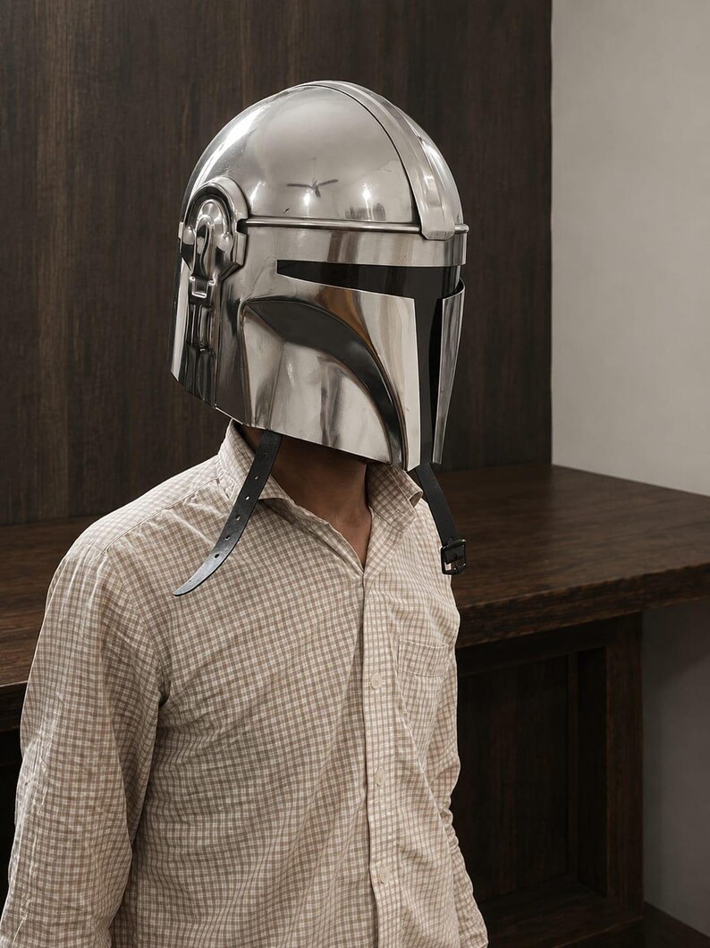 May include: A polished silver-colored metal helmet with a smooth, reflective surface and a T-shaped black visor opening. The helmet features a central ridge, side ear-cap details, and two thin black leather chin straps hanging down from the sides.
