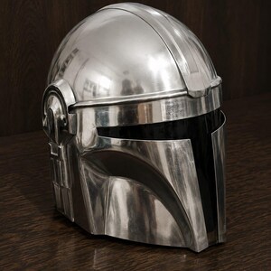 May include: A polished silver-colored helmet with a smooth, reflective metallic finish. The helmet features a T-shaped visor opening with a dark, opaque black face shield and distinct structural paneling along the sides and top dome section.