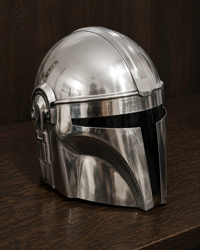 May include: A polished silver-colored helmet with a smooth, reflective metallic finish. The helmet features a T-shaped visor opening with a dark, opaque black face shield and distinct structural paneling along the sides and top dome section.