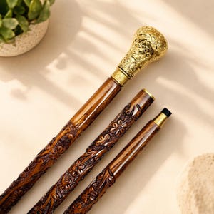 Pode incluir: Uma bengala dobrável composta por três segmentos de madeira castanha escura com entalhes florais complexos. O punho superior é feito de metal dourado polido com padrões em relevo, e as juntas apresentam ponteiras de metal dourado.