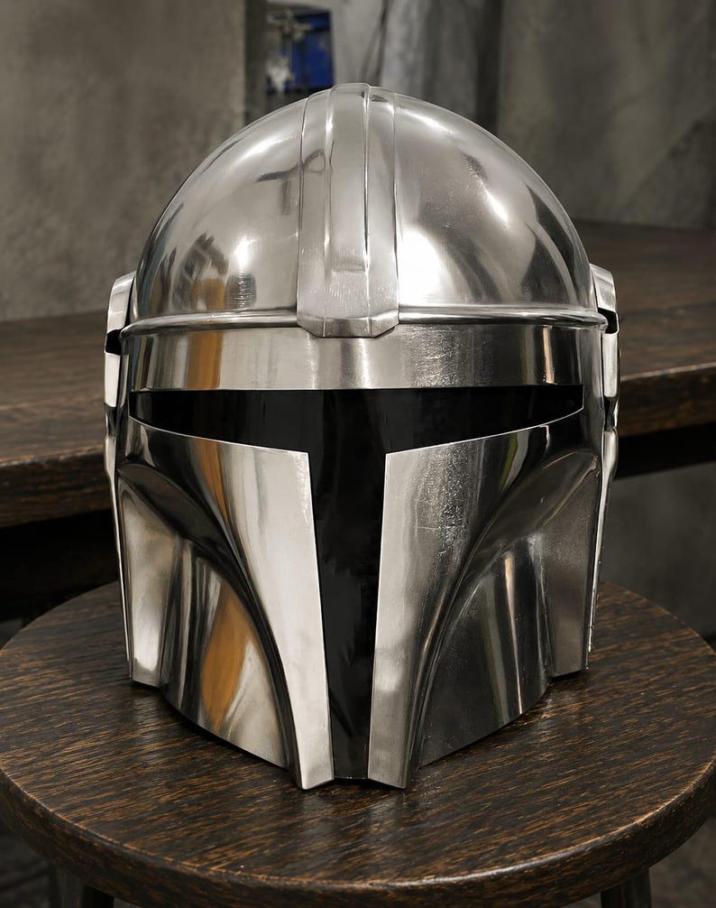 May include: A polished silver-colored metal helmet featuring a smooth, rounded dome, a central vertical ridge, and a wide, horizontal black visor opening. The helmet has a distinct T-shaped faceplate design with curved cheek sections and side panels.