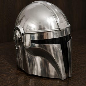 May include: A polished silver-colored helmet with a smooth, reflective metallic finish. The helmet features a T-shaped visor opening with a dark, opaque black face shield and distinct structural paneling along the sides and top dome section.