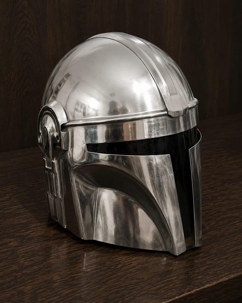 May include: A polished silver-colored helmet with a smooth, reflective metallic finish. The helmet features a T-shaped visor opening with a dark, opaque black face shield and distinct structural paneling along the sides and top dome section.