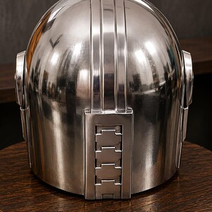 May include: A polished silver-colored helmet with a smooth, reflective dome surface. The helmet features a vertical, rectangular panel on the front center with four horizontal, recessed rectangular segments and circular side attachments.