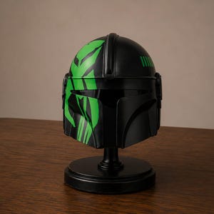 May include: A matte black helmet featuring a T-shaped visor and a prominent bright green graphic design on the left side. The helmet is mounted on a circular black display stand, showcasing a smooth finish and distinct angular structural details.