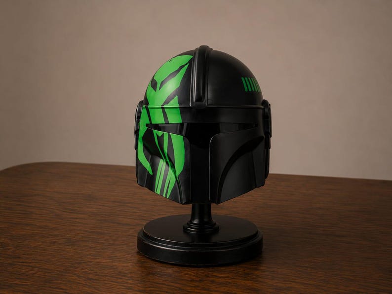 May include: A matte black helmet featuring a T-shaped visor and a prominent bright green graphic design on the left side. The helmet is mounted on a circular black display stand, showcasing a smooth finish and distinct angular structural details.
