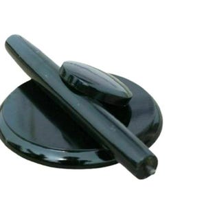 May include: A glossy black circular base with a smooth, rounded surface, featuring a long, cylindrical black handle positioned diagonally across the top and a small, oval-shaped black accent piece resting near the center of the base.