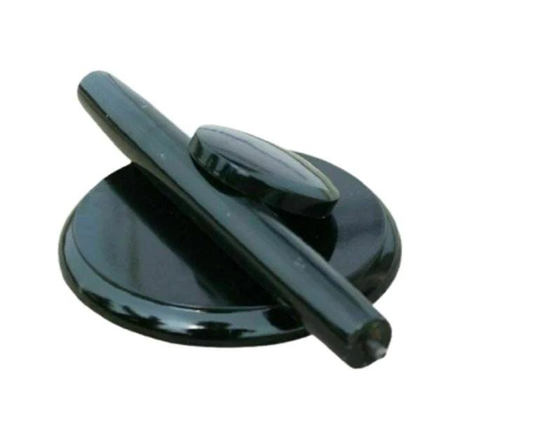 May include: A glossy black circular base with a smooth, rounded surface, featuring a long, cylindrical black handle positioned diagonally across the top and a small, oval-shaped black accent piece resting near the center of the base.