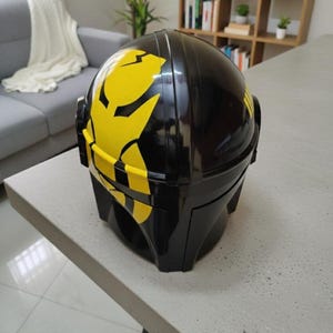 May include: A glossy black helmet featuring a prominent yellow graphic design on the side. The helmet has a smooth, rounded dome shape with a structured visor area and side panels, showcasing a bold contrast between the black and yellow colors.
