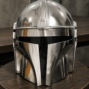 May include: A polished silver-colored metal helmet featuring a smooth, rounded dome, a central vertical ridge, and a wide, horizontal black visor opening. The helmet has a distinct T-shaped faceplate design with curved cheek sections and side panels.