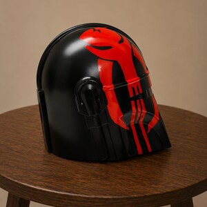 May include: A black helmet featuring a glossy finish and a prominent red graphic design on the front. The design includes a stylized emblem on the forehead area and vertical red stripes extending down the faceplate, with a dark visor section.