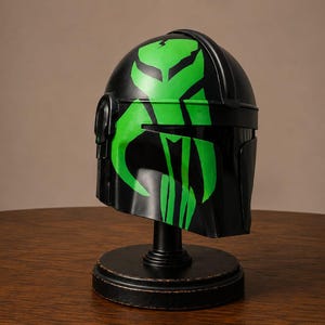 May include: A black helmet featuring a prominent, bright green graphic design of a stylized skull emblem on the front. The helmet is mounted on a circular black stand, showcasing a smooth, glossy finish and a T-shaped visor opening.