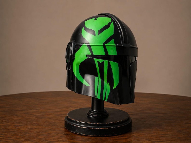 May include: A black helmet featuring a prominent, bright green graphic design of a stylized skull emblem on the front. The helmet is mounted on a circular black stand, showcasing a smooth, glossy finish and a T-shaped visor opening.