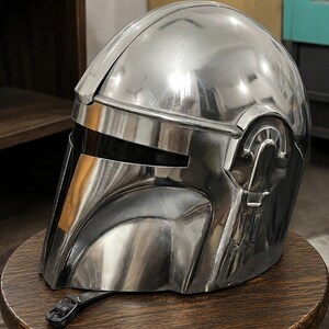 May include: A polished silver-colored helmet with a smooth, reflective metallic finish. The helmet features a T-shaped visor opening with a dark, opaque faceplate, a prominent central ridge, and a black chin strap with a buckle attached at the base.