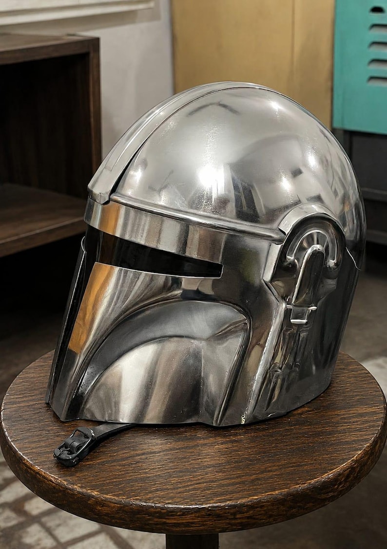 May include: A polished silver-colored helmet with a smooth, reflective metallic finish. The helmet features a T-shaped visor opening with a dark, opaque faceplate, a prominent central ridge, and a black chin strap with a buckle attached at the base.