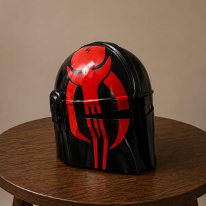 May include: A glossy black helmet featuring a prominent red graphic design on the front. The design depicts a stylized skull-like emblem with sharp, curved lines and a small lightning bolt symbol at the top center of the forehead area.