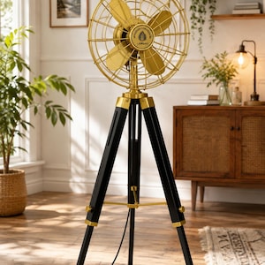 May include: A floor fan featuring a gold-coloured metal cage and four blades, mounted on a black wooden tripod stand with gold accents. The centre of the fan head displays a circular logo with the text LAWANY and a leaf icon in the middle.
