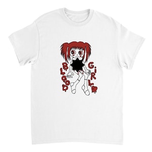 Puede incluir: Camiseta blanca con una ilustración de dibujos animados de una muñeca. La muñeca tiene el pelo rojo y ojos grandes, vestida con un vestido negro. Las palabras "BLOOD GIRL" están impresas en rojo, con letras goteando.