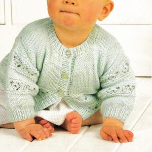Op de afbeelding: Een baby draagt een lichtgroen gebreid vest met drie knopen. Het vest heeft decoratieve ajourdetails op de mouwen. De baby zit op een witte ondergrond.