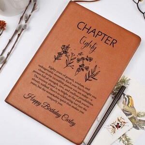 Puede incluir: Un diario de piel sintética color canela con el texto "CHAPTER Eighty" y "Happy Birthday Carley", una ilustración floral y un párrafo de texto. El diario se acompaña de un bolígrafo negro, una taza de café blanca y gafas negras.
