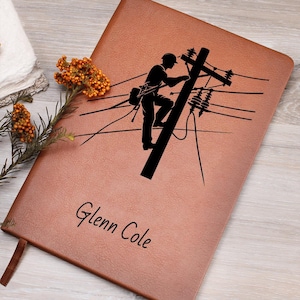 May include: A tan leather-like notebook featuring a black graphic of a utility worker climbing a power pole. The name Glenn Cole is printed in black script below the graphic. The cover has a smooth texture and a rectangular shape.