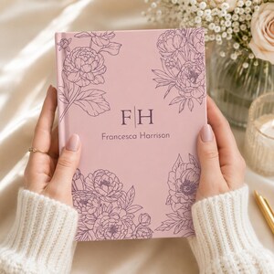 Puede incluir: Un diario de tapa dura de color rosa claro con dibujos de líneas de color morado oscuro de flores de peonía en las esquinas. El centro muestra las iniciales FH separadas por una línea vertical, con el nombre Francesca Harrison debajo.