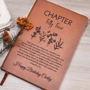 May include: A tan leather-like journal featuring the text "CHAPTER Fifty Four" and a floral line art illustration. Below, a printed message reads "Fifty Four years of growth, wisdom, and unforgettable moments." It ends with "Happy Birthday Carley."
