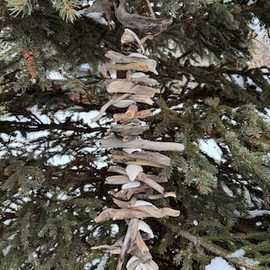 Può includere: Una carillon di legno di deriva fatto a mano appeso a un pino. La carillon è composta da legno stagionato di varie dimensioni e forme, con piccole pietre bianche intercalate. I toni naturali del legno contrastano con il verde dell'albero e la neve.