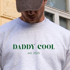 Könnte beinhalten: Ein hellgraues Sweatshirt mit dem grünen Text "DADDY COOL est. 2025". Die Person auf dem Bild trägt eine braune Baseballkappe mit dem Text "La Poésie PARIS". Das Sweatshirt hat einen Rundhalsausschnitt.