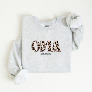 Könnte beinhalten: Hellgraues Sweatshirt mit dem Wort "OMA" in einem Leopardenmuster. Unter dem Wort "OMA" steht der Text "est. 2025". Das Sweatshirt hat einen Rundhalsausschnitt und lange Ärmel.