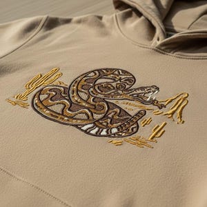 May include: A tan hoodie featuring an embroidered snake design in shades of brown, tan, and white. The snake is coiled with its mouth open, surrounded by embroidered cacti. The hoodie has a drawstring hood and a kangaroo pocket.