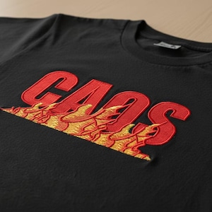 Op de afbeelding: Zwart T-shirt met het woord "CAOS" geborduurd in rood, met gele en oranje vlammen eronder. Het shirt heeft een ronde hals en korte mouwen.