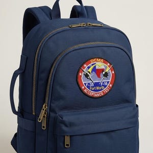 May include: Navy blue backpack with a patch featuring the text "DCMA Acceptance Test" and images of F-35 and F-16 fighter jets. The backpack has multiple zippered compartments and adjustable straps.