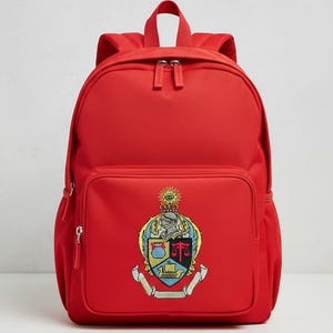 Może przedstawiać: Jasnoczerwony plecak z przednią kieszenią i górnym uchwytem. Plecak posiada kolorowy, haftowany herb na przedniej kieszeni. Herb zawiera tarczę, koronę i inne symbole. Plecak posiada dwa zamki błyskawiczne w głównej komorze.