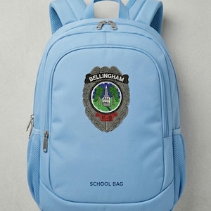 May include: A light blue school backpack featuring a Bellingham emblem and the words "SCHOOL BAG". The emblem includes a building, trees, and the text "K-9". The backpack has mesh side pockets and a top handle.