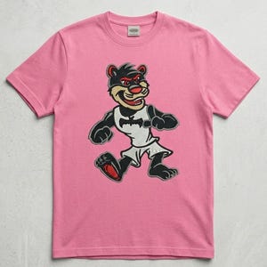 Puede incluir: Una camiseta rosa con un personaje de dibujos animados en blanco y negro, con detalles rojos. El personaje lleva una camiseta de baloncesto blanca con un logotipo de murciélago. La camiseta es de tejido suave.