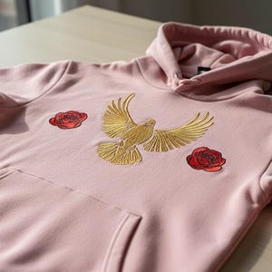 Puede incluir: Una sudadera con capucha rosa claro con bolsillo de canguro y capucha. Presenta una paloma dorada bordada con las alas extendidas, flanqueada por dos parches de rosas rojas. La sudadera está hecha de un material suave.