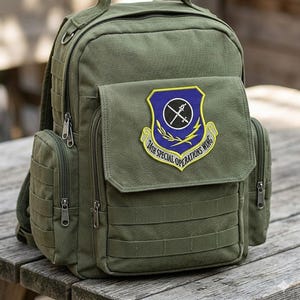 May include: Olive green backpack with multiple zippered compartments and a top handle. The front flap features a patch with a blue and gold emblem and the text "24th Special Operations Wing". The backpack has adjustable straps and a tactical design.