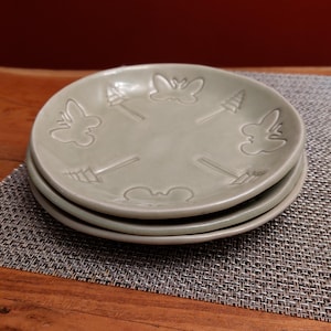 Peut inclure: Trois assiettes en céramique vert clair empilées. Chaque assiette présente un motif en relief d'un papillon, d'une flèche et d'un arbre. Les assiettes sont empilées sur un set de table gris, avec une surface en bois au premier plan.