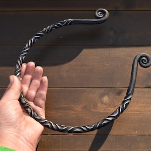 May include: A hand-forged, wrought iron towel ring. The dark metal ring features a twisted design along the curved bar and decorative spiral ends. The towel ring is shown against a wooden background.