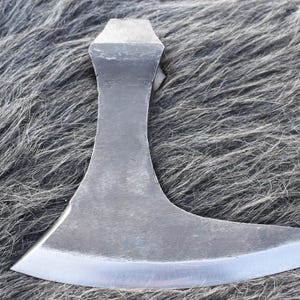 Op de afbeelding: Een handgesmede stalen bijlkop met een brede, gebogen snijrand en een taps toelopende achterkant. Het metalen oppervlak heeft een donkergrijze, matte afwerking met een gepolijste, reflecterende rand langs het blad, wat een functionele vorm toont.