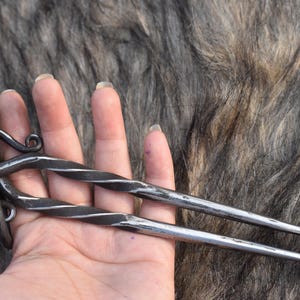 Peut inclure: Épingles à cheveux en métal forgé à la main avec un design torsadé. Les accessoires pour cheveux sont gris foncé avec des accents argentés et un dessus bouclé. Les extrémités pointues sont visibles. Le fond est texturé, brun et gris.