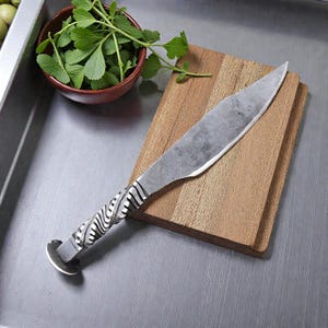 Può includere: Un coltello in acciaio forgiato a mano con una lama strutturata e un manico decorativo. L'impugnatura presenta un design in metallo attorcigliato con sezioni chiare e scure alternate. Il coltello è appoggiato su un tagliere di legno, con una piccola ciotola di erbe verdi nelle vicinanze.