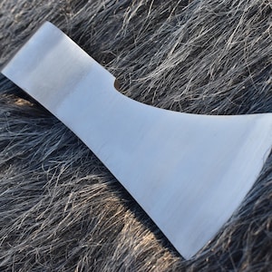 Op de afbeelding: Een zilverkleurige bijlkop met een gebogen blad en een platte bovenkant. De bijlkop ligt op een getextureerd, donkergrijs oppervlak, mogelijk vacht of stof. Het blad lijkt scherp en klaar voor gebruik.