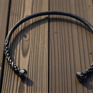 May include: A dark gray metal torc necklace. The necklace has a circular design with decorative, twisted ends and rounded terminals. The necklace is displayed on a wooden surface.