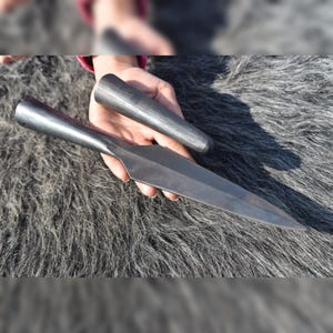 May include: A dark gray metal spearhead with a pointed tip and a hollow cylindrical socket base, accompanied by a separate, smaller, solid dark gray metal cylindrical component, both featuring a smooth, matte, forged finish.