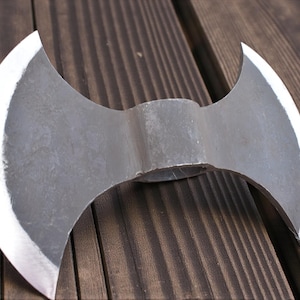 May include: A double-bladed axe with a silver-colored head and a dark gray body. The axe is placed on a wooden surface with a dark brown color.