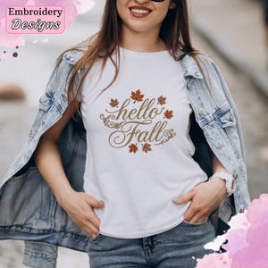 Puede incluir: Una camiseta blanca con el texto bordado en marrón hello Fall rodeado de pequeñas hojas de arce marrones. La camiseta se lleva debajo de una chaqueta vaquera azul claro. El diseño está centrado en el pecho de la prenda.